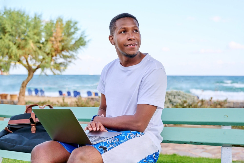 A man sitting on a sandy beach, engrossed in his work, with a laptop open in front of him. The tranquil ocean waves and blue sky create a serene backdrop as he embraces hybrid working, blending productivity and relaxation in a picturesque setting.